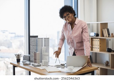 Confident African American female architect measures building model dimensions using compass in modern office. Conceptualizes urban architecture project with enthusiasm and focus.
