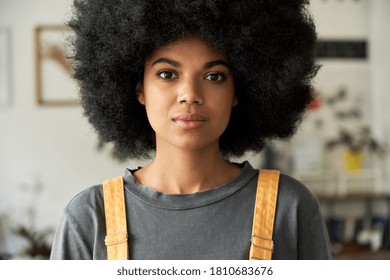 Confident African American gen z hipster female student with Afro hair looking at camera standing indoors, at home, in modern creative office. Mixed race young woman close up headshot portrait.