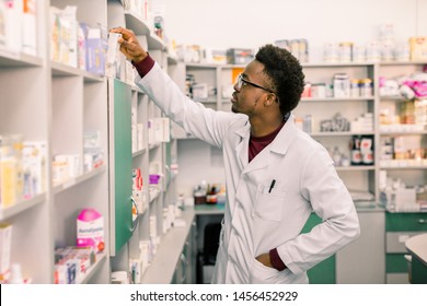 Confident African American Man Pharmacist Standing In Interior Of Pharmacy And Searching The Drug On Shelves In Pharmacy