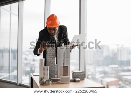 Confident african american man in suit using laptop looking at the architecture design of buildings at desk with residential project maquette at modern office with panoramic city view