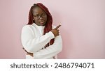 Confident african american woman with braids wearing glasses and white sweater over pink background pointing sideways