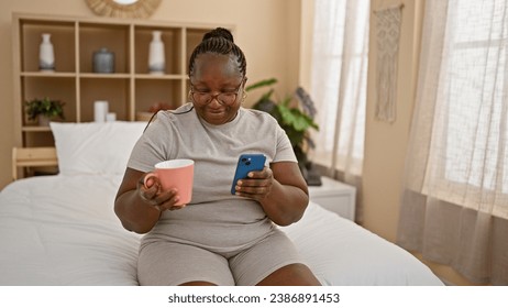 Confident african american woman comfortable in her skin, relaxing in bed, drinking morning coffee, texting on phone, braids resting on the pillow, her smile lighting up the room. - Powered by Shutterstock