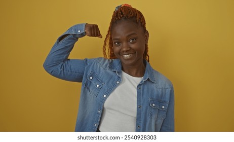 A confident african american woman in a denim jacket poses with a flexed arm against a yellow background. - Powered by Shutterstock