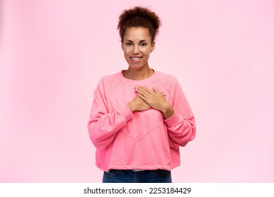Confident African American woman in pink sweatshirt, holding hands at heart level, smiling looking at camera isolated on pink background. Breast Cancer Awareness Day. Cancer Campaign. Pink October 1st