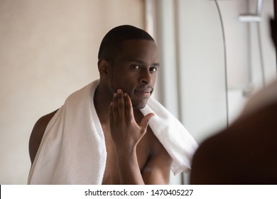 Confident african american young man standing in bathroom after shower with towel on shoulder, looking in mirror, touching soft skin after shave, applying lotion. Skincare, morning hygiene concept.