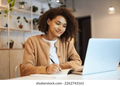 Confident African American young woman with stylish afro hairstyle, smart student studying, taking notes on notepad watching training courses on laptop. Online business or education concept
