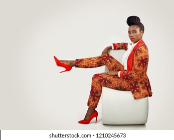 Confident African business woman  wearing an African print suit and heels with a sophisticated hair style sitting in a clean space with her leg propped up.
