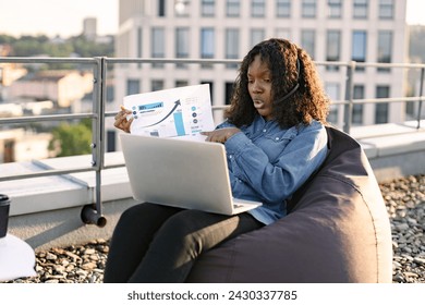 Confident African woman in headset maintaining video communication via modern gadgets in sunlight on rooftop. Efficient adult worker conducting online data analysis via bar graphs in outdoor office. - Powered by Shutterstock