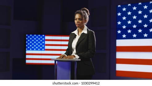 Confident African-American woman holding press conference while standing alone on podium with background of American flag on screen. - Powered by Shutterstock