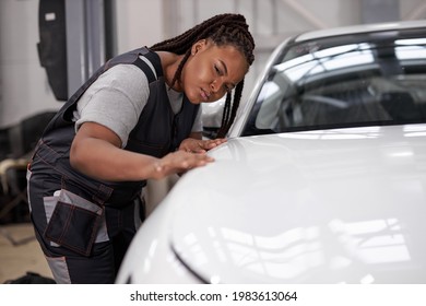 Confident Afro American Female Auto Mechanic Touching Surface Of White Repainted And Clean Car Body In Auto Repair Shop. Pretty Black Woman Mechanic In Uniform Working In Workshop, Looking Serious
