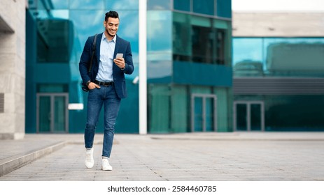 Confident arabic guy programmer with backpack walking by street, checking his mobile phone, chatting with friends, going home from office, panorama with copy space, full length