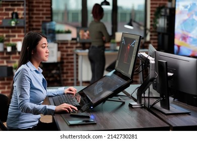 Confident asian 3D model creator sitting at desk with multiple monitors while working on CGI. Digital artist creating computer generated imagery for game development while in creative agency workspace - Shutterstock ID 2142730281