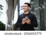 Confident Asian businessman in modern city, smiling while using smartphone outdoors, wearing professional suit, representing success and connectivity in digital age.