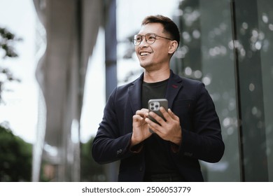 Confident Asian businessman in modern city, smiling while using smartphone outdoors, wearing professional suit, representing success and connectivity in digital age. - Powered by Shutterstock