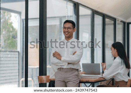 Confident asian businessman professional standing with arms crossed with female colleague working in background. Leadership and business confidence concept.