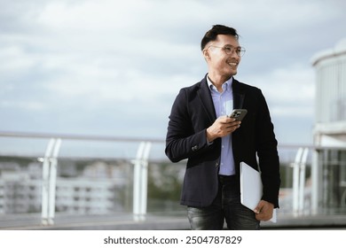 A confident Asian businessman standing in a modern city, smiling while holding a smartphone, dressed in a sharp suit and glasses, embodying corporate success and professionalism. - Powered by Shutterstock