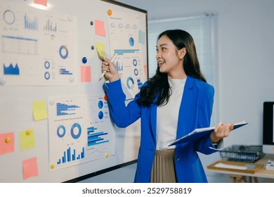 Confident asian businesswoman in bright office, smiling, pointing at whiteboard with sticky notes and graphs, explaining business plan to team during meeting - Powered by Shutterstock