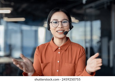 A confident Asian businesswoman with a headset leading a virtual meeting in a modern office environment. She exhibits a friendly smile and open hand gestures. - Powered by Shutterstock