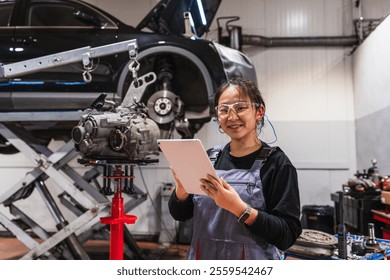 Confident asian female mechanic using a digital tablet, with a car lift and gearbox in the background, working in a professional vehicle repair shop - Powered by Shutterstock