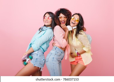 Confident Asian Girl In Denim Shorts Posing With International Female Friends On Indoor Photoshoot. Beautiful Ladies In Bright Clothes Dancing With Pleasure And Laughing Together.