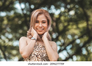 A Confident Asian Lady With Blonde Dyed Hair In A Leopard Top. Outdoor Park Scene.