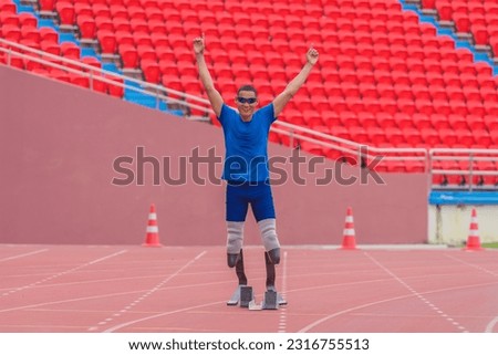 confident asian male athlete, equipped with two prosthetic running blades, alertly prepares to position himself at start of speed run on sports stadium track, embodying strength and anticipation