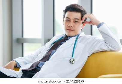 Confident Asian Male Physician In White Gown And With Stethoscope Sitting On Sofa And Leaning On Hand In Hospital While Looking At Camera