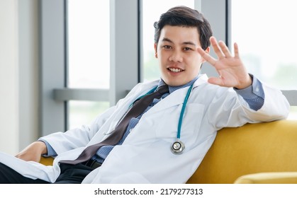 Confident Asian Male Physician In White Gown And With Stethoscope Sitting On Sofa And Greeting In Hospital While Looking At Camera