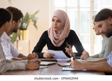 Confident Asian muslim female team leader holding corporate meeting, serious businesswoman wearing hijab holding document, discussing project statistics with colleagues, sitting at table in boardroom