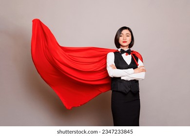 Confident asian waitress standing with folded arms and posing in fluttering superman red cape. Young attractive woman receptionist dressed in hero cloak and catering service uniform
