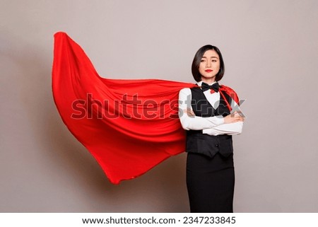 Confident asian waitress standing with folded arms and posing in fluttering superman red cape. Young attractive woman receptionist dressed in hero cloak and catering service uniform