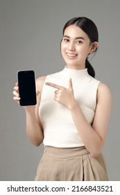 Confident Asian Woman Holding Phone Show Blank Screen And Looking At Camera Isolated On Gray Background. Portrait Of A Beautiful Girl In The Studio.