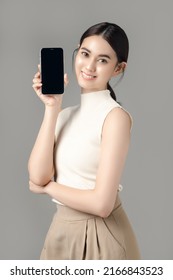 Confident Asian Woman Holding Phone Show Blank Screen And Looking At Camera Isolated On Gray Background. Portrait Of A Beautiful Girl In The Studio.