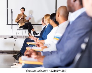 Confident Asian Woman Sitting On Conference Room Stage With Microphone In Hand, Lecturing During Business Seminar