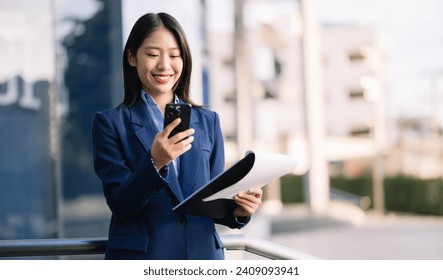 Confident Asian woman with a smile standing holding notepad and tablet at out side office. big city on business center background. - Powered by Shutterstock