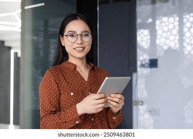 Confident Asian worker in modern office environment holding tablet. Dressed in polka dot blouse , she exhibits friendly and professional demeanor, representing diverse workforce in corporate setting - Powered by Shutterstock