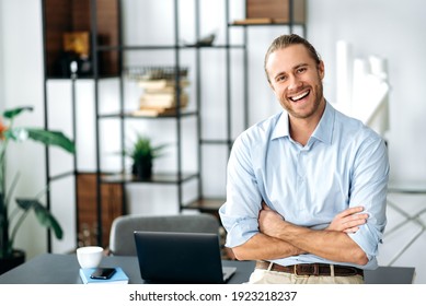 Confident attractive caucasian stylishly dressed guy, ceo, freelancer or employee, standing near his desk, with arms crossed, looking at the camera and smiling friendly