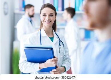 Confident Attractive Female Doctor In The Office Holding Medical Records And Folders, She Is Smiling At Camera, Medical Staff On The Background