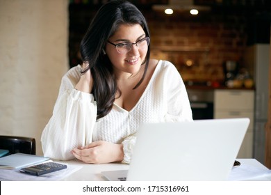 Confident attractive young plus size female in white blouse using laptop for distant work, sitting in home interior. Chubby student girl doing homework, surfing internet on portable computer - Powered by Shutterstock