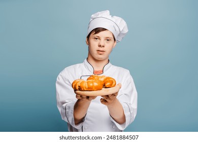 Confident baker holding freshly baked croissants on wooden plate standing isolated on blue background looking at camera. Smiling teenager in uniform. Food concept - Powered by Shutterstock