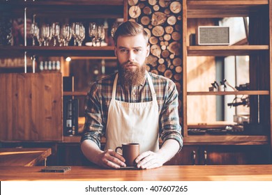 Confident Barista. Young Bearded Man In Apron Looking At Camera And Holding Coffee Cup While Standing At Bar Counter