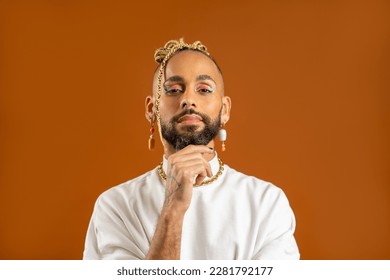 Confident bearded black gay man with bright makeup standing isolated on orange background, dressed white. Exudes sense of pride and individuality. Diversity power of personal style