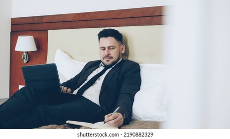 Confident Bearded Businessman Lying On Bed Using Laptop Computer And Writing Notes At Notepad While Working In Hotel Room. Travel, Business And People Concept