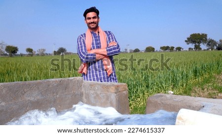 A confident bearded farmer standing near a tubewell with his folded arms - leisure time, relaxation. A happy worker laborer in casual clothes and wearing a Safa posing for the camera in his green...