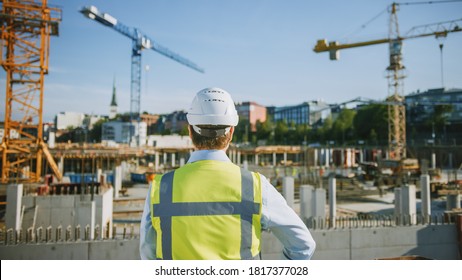 Confident Bearded Head Civil Engineer-Architect In Sunglasses Is Standing Outside With His Back To Camera In A Construction Site On A Bright Day. Man Is Wearing A Hard Hat, Shirt And A Safety Vest.