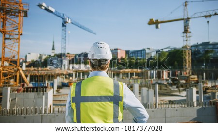 Confident Bearded Head Civil Engineer-Architect in Sunglasses is Standing Outside with His Back to Camera in a Construction Site on a Bright Day. Man is Wearing a Hard Hat, Shirt and a Safety Vest.