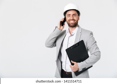 Confident bearded man builder wearing suit and hardhat standing isolated over white background, talking on mobile phone