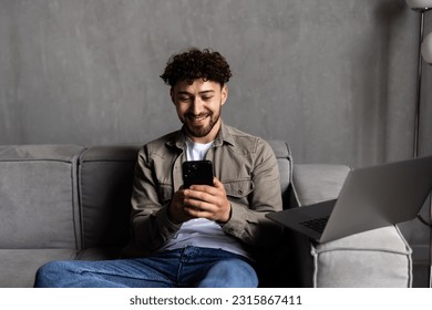 Confident bearded man is sitting on couch and typing on smartphone.