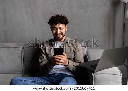 Confident bearded man is sitting on couch and typing on smartphone.