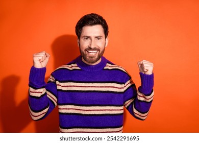 Confident bearded man in striped sweater celebrating success against vibrant orange background - Powered by Shutterstock